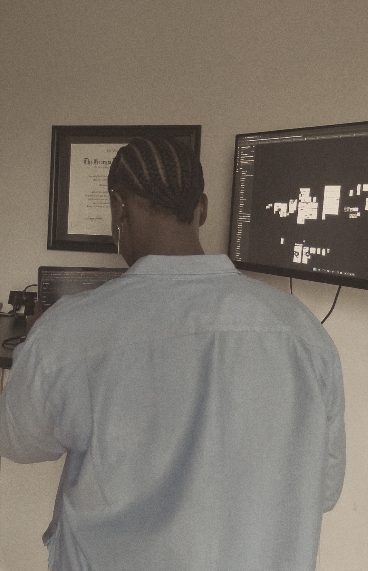 Khadijat Amoo working at a desk, viewed from behind, with a Georgia Tech diploma and design work on a monitor in the background